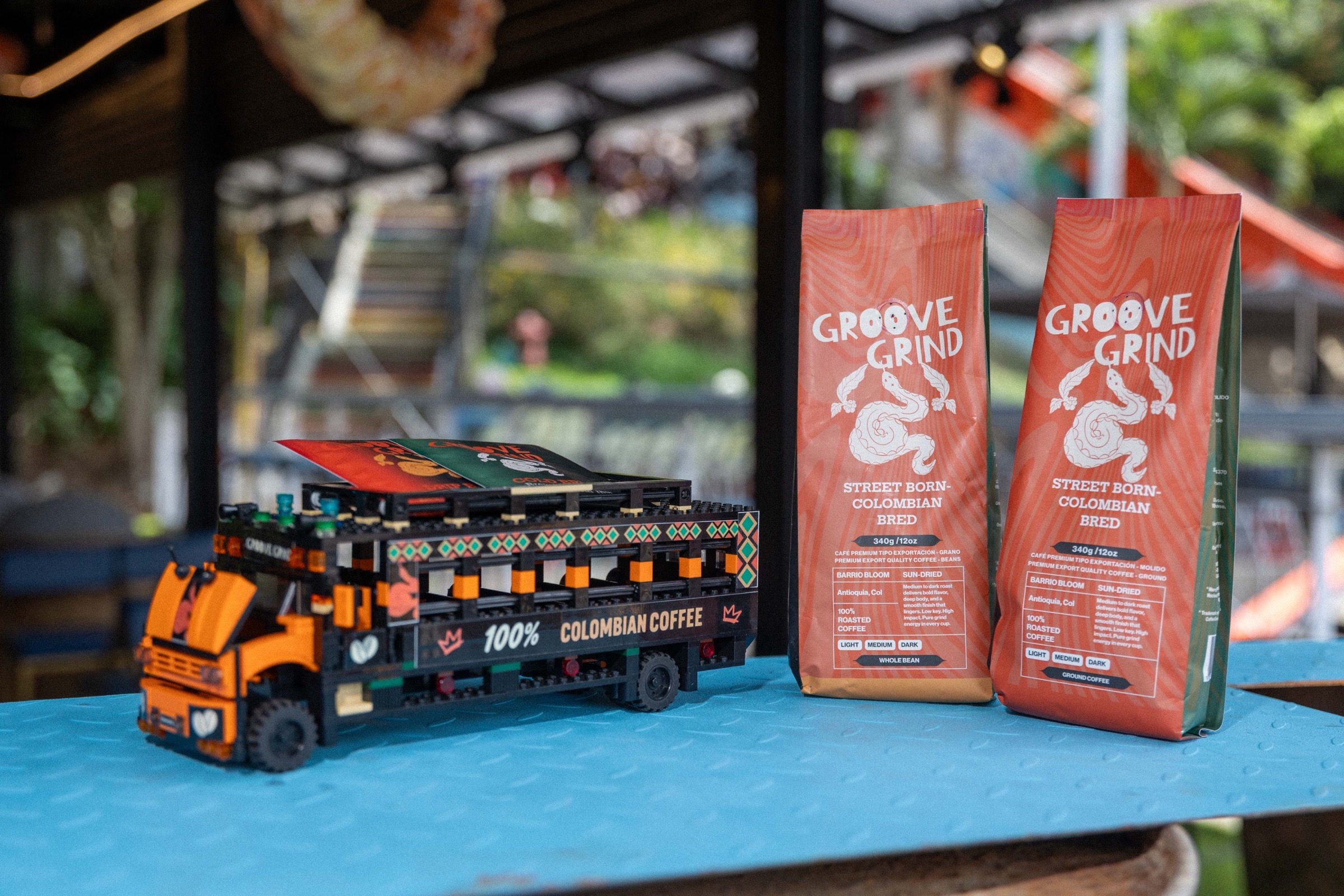 A wooden model of a Colombian chiva painted with 100% Colombian Coffee, beside two Groove Grind bags — whole bean and ground — on a blue table in Comuna 13.