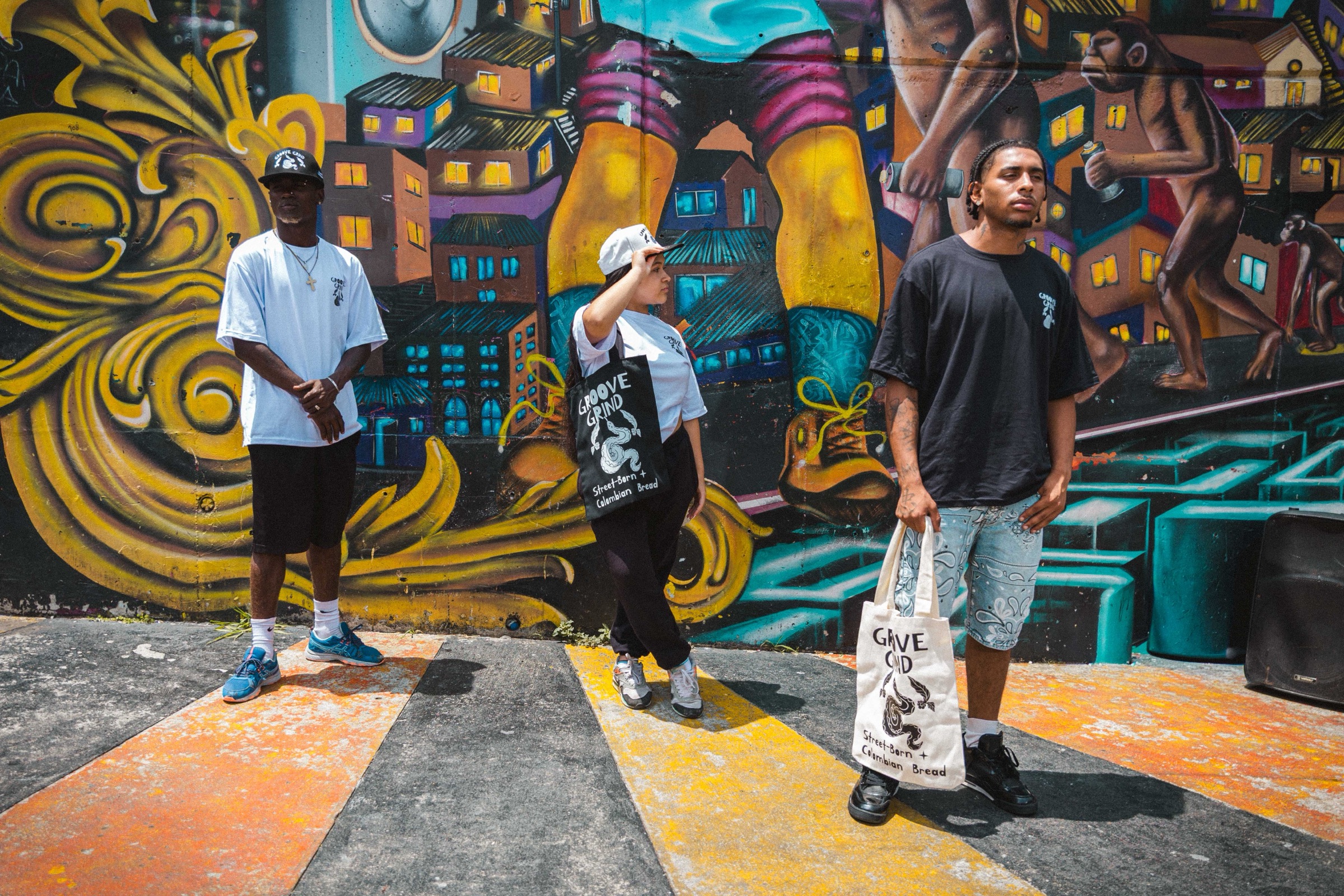 Three people wearing Groove Grind shirts and totes stand on a painted crosswalk in front of a Comuna 13 mural.